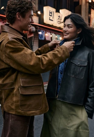 Man in brown suede jacket smiling at a woman in a black jacket adjusting her collar outdoors at night.