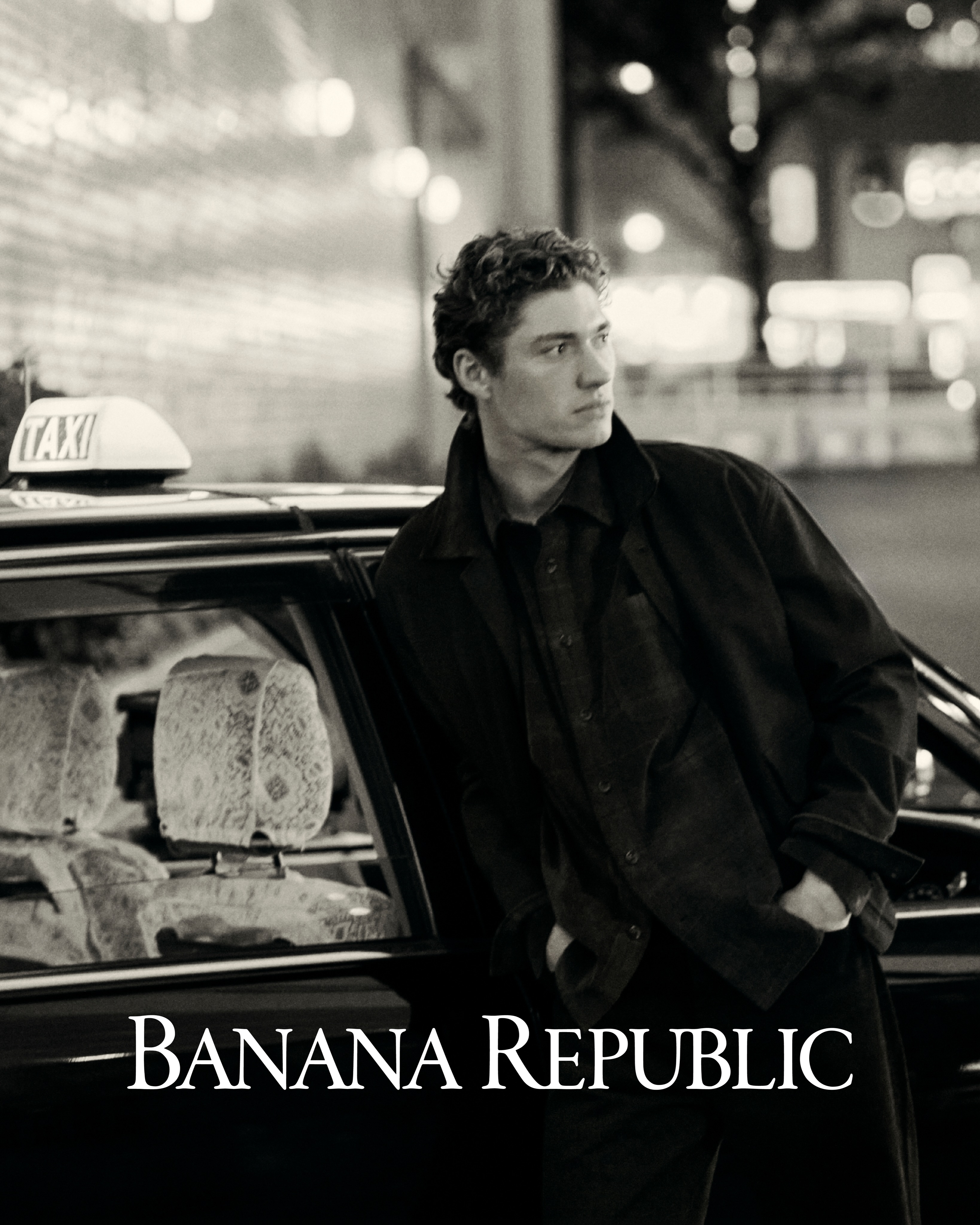 Man wearing a dark jacket leaning against a taxi cab at night with Banana Republic text overlay.