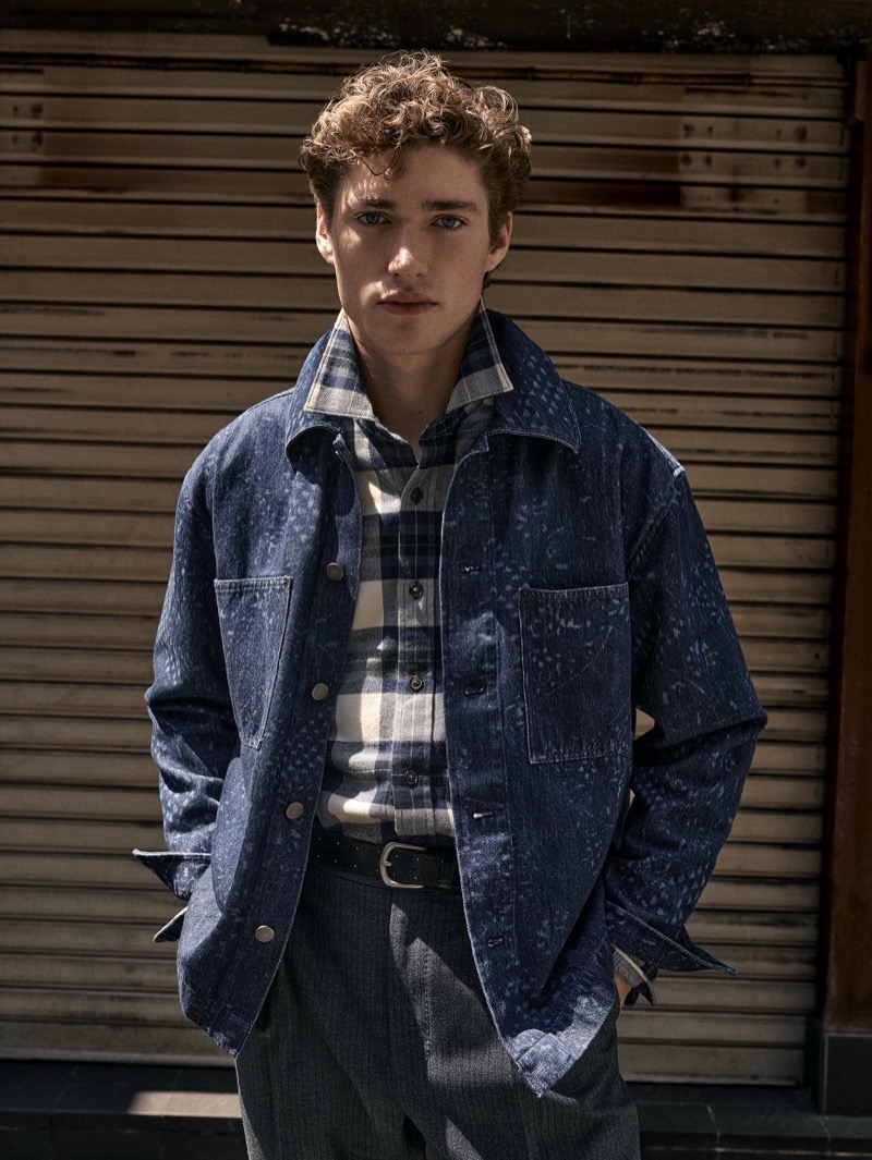 A man in a blue patterned denim jacket and plaid shirt posing against a slatted metallic shutter for Banana Republic.