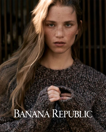 Portrait of a woman with long blonde hair wearing a chunky brown knit sweater against a dark slatted background.