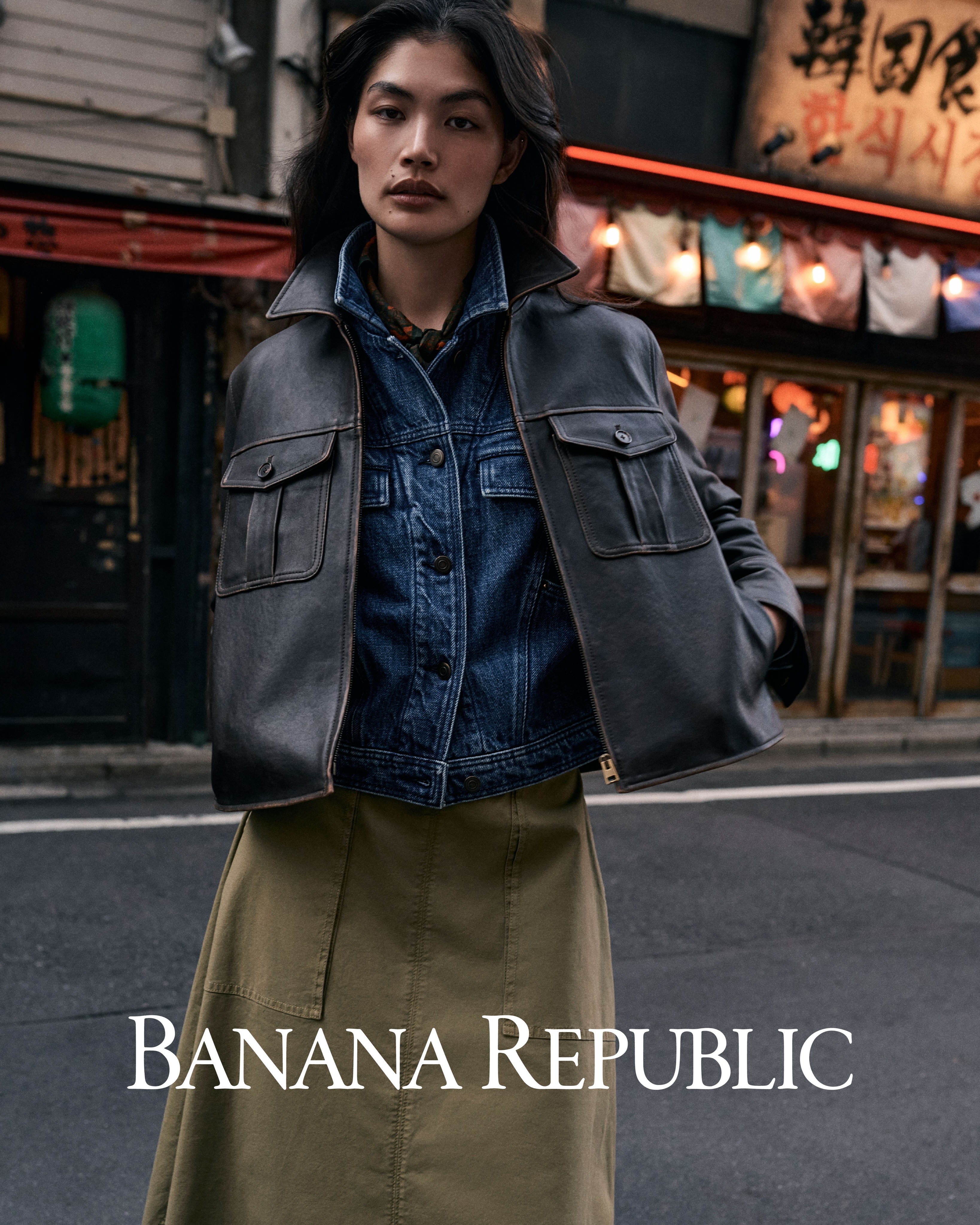 Model wearing a dark leather jacket over a denim jacket standing on a street with storefronts in the background.