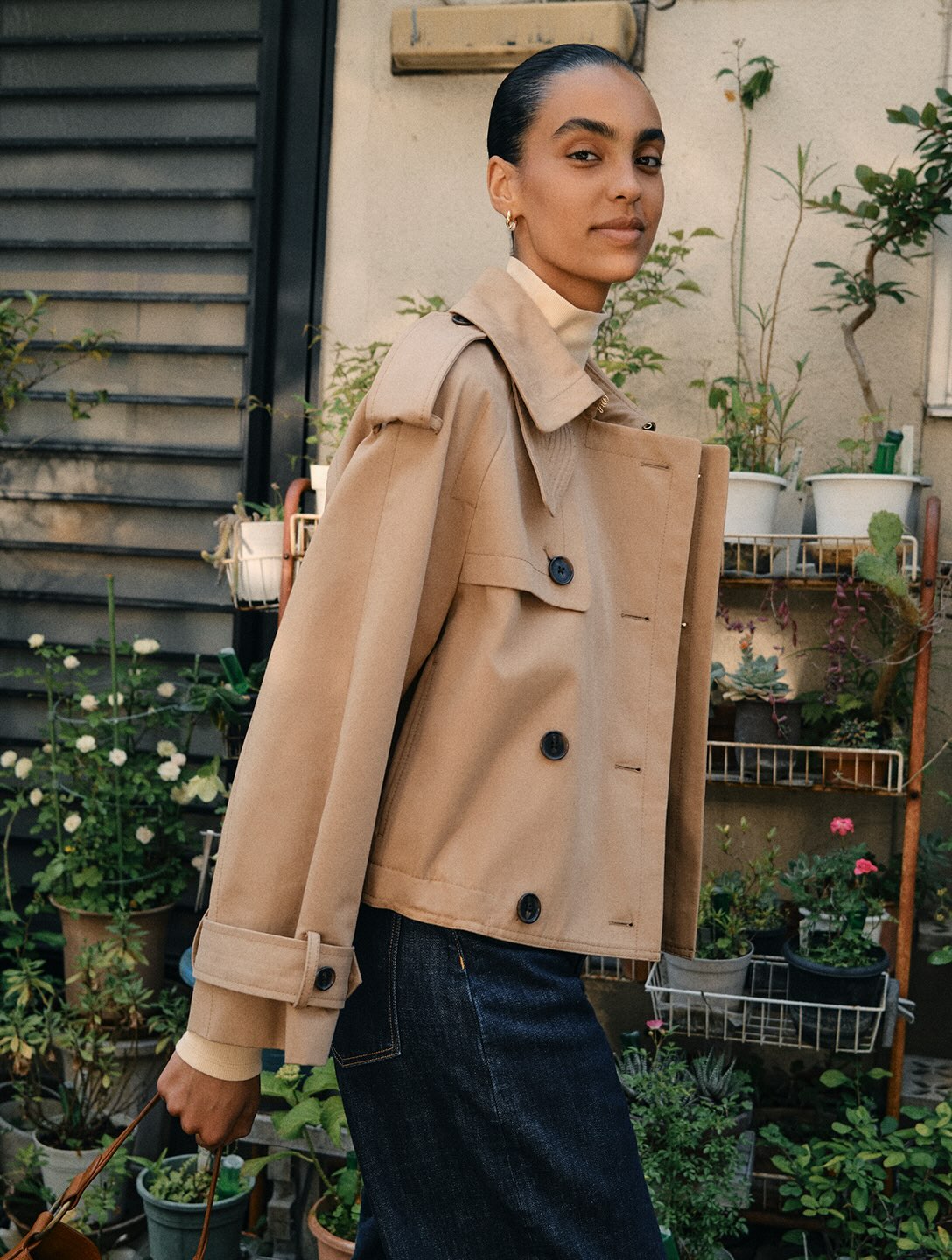 Model in camel jacket and dark denim jeans posing outdoors beside potted plants