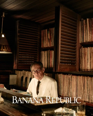 An elderly man in a white shirt and tie stands behind a bar with shelves filled with records in a warm, dimly lit lounge setting.