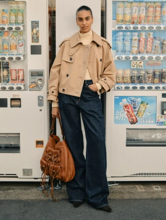 Model standing between two vending machines wearing a tan trench jacket, turtleneck, and dark jeans while holding a brown leather bag
