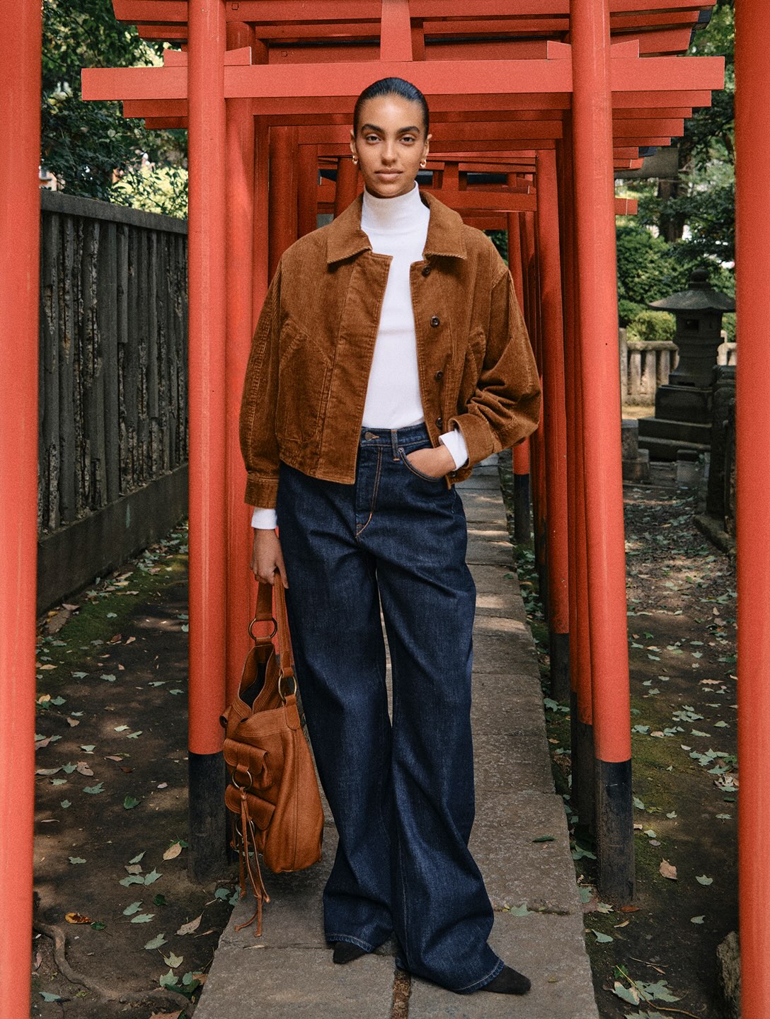 Model standing on a stone pathway framed by orange torii gates wearing a brown jacket, white turtleneck, and blue jeans
