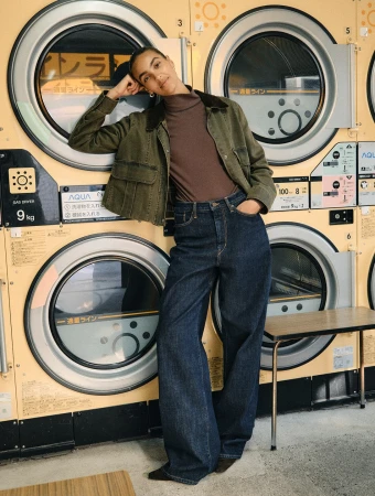 Woman in an olive green jacket, brown turtleneck, and dark denim jeans posing against industrial washing machines