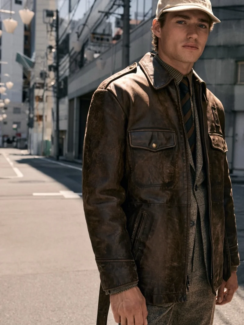 A man wearing a brown leather jacket and a baseball cap standing on a city street.