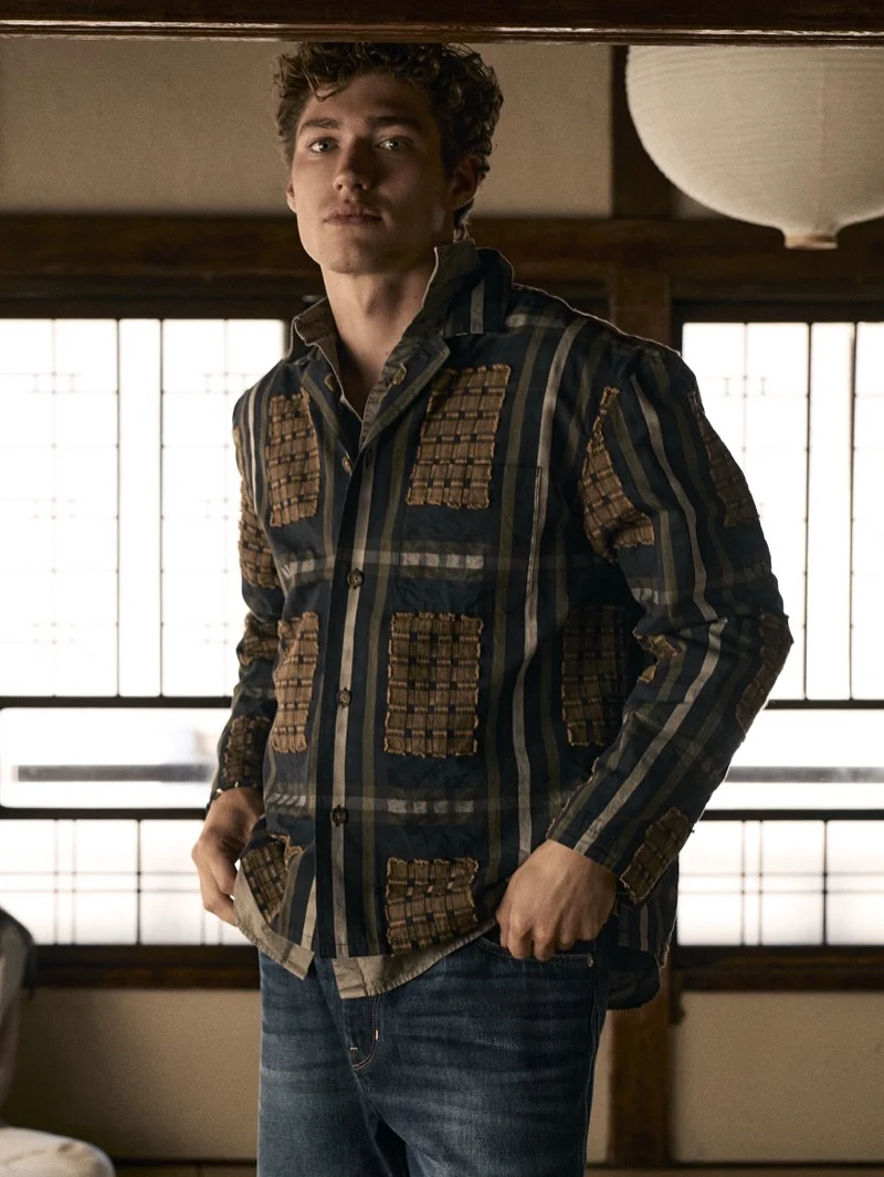 Young man wearing a patterned shirt and blue jeans standing indoors before a window panel with a paper lantern hanging above.