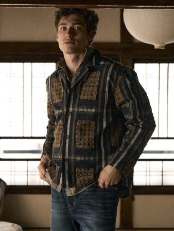 Young man wearing a patterned shirt and blue jeans standing indoors before a window panel with a paper lantern hanging above.