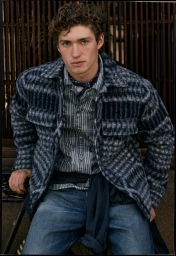 Young man with curly hair wearing a plaid overshirt, striped shirt, and blue jeans against a slated wooden background