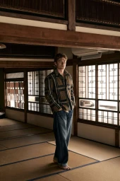 A man wearing a patterned long-sleeve shirt and jeans standing barefoot on tatami mats inside a traditional interior.