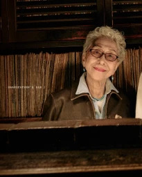 Elderly woman with short curly grey hair wearing glasses and a leather jacket seated in a dimly lit bar.