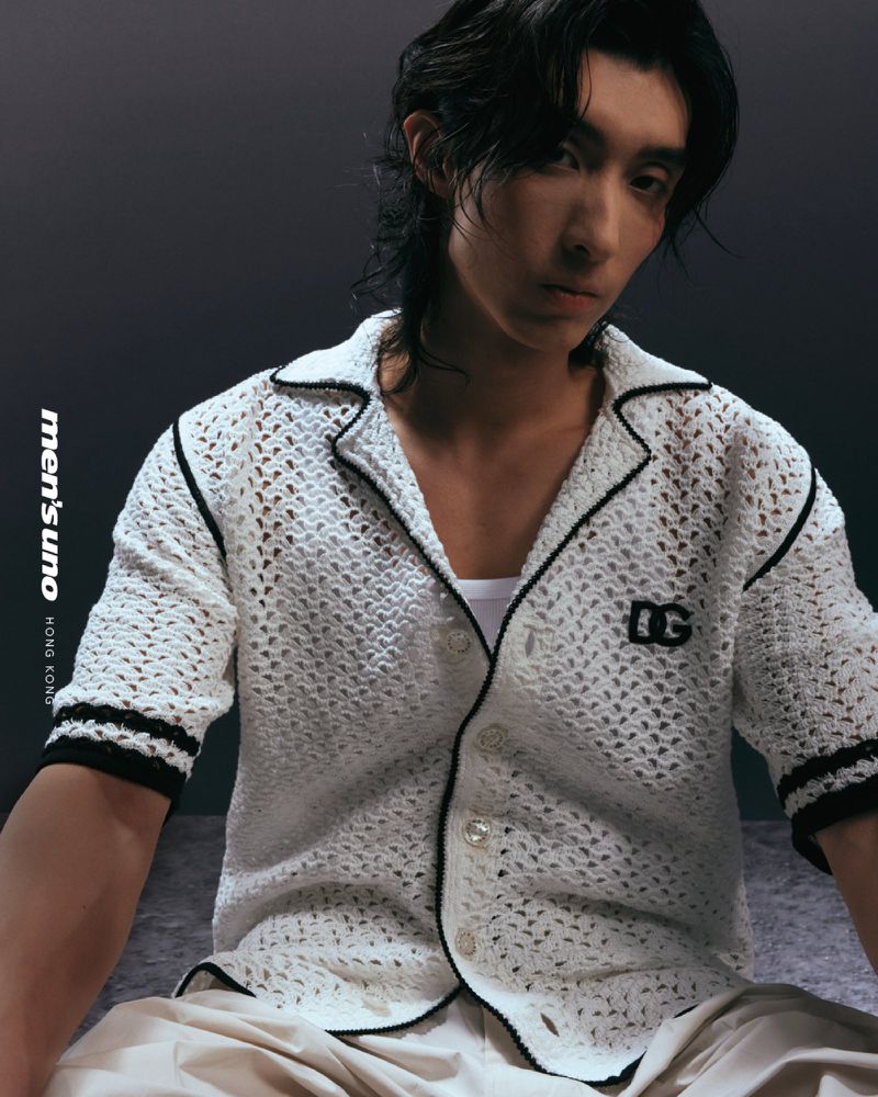 Portrait of an Asian man wearing a white crocheted shirt and tan pants against a dark gray background in a fashion editorial.