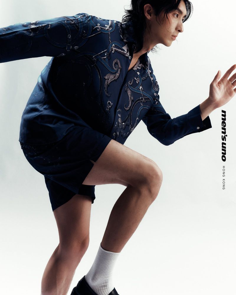 Editorial fashion shot of an Asian male model in a navy, decorative jacket and black shorts, posed dynamically against a white backdrop.