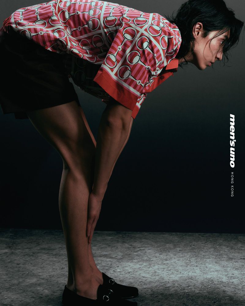 Asian male fashion model with short hair wearing a patterned shirt and shorts, posing in an indoor photo studio setting with dark background.