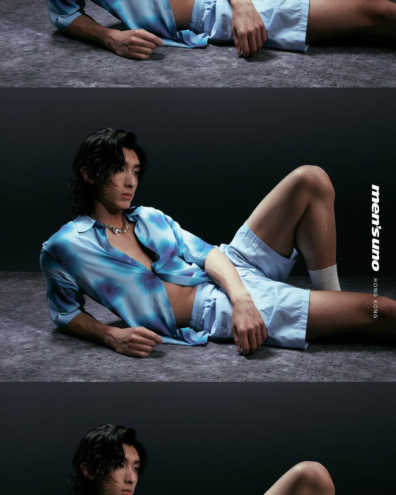 Adult male fashion model poses while laying down in a blue tie-dye shirt and shorts against a dark grey textured background in studio lighting.