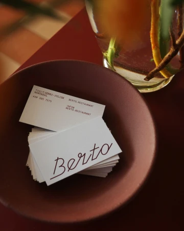 Stack of white business cards with the name Berto printed in red script resting inside a deep red ceramic bowl.