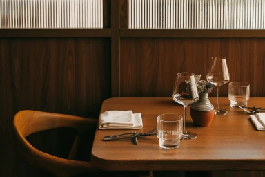 Set wooden table with wine glasses, ribbed tumblers, and a ceramic vase against dark wood panels and reeded glass.