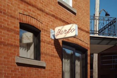 Exterior brick building wall featuring a light-colored projecting sign with red cursive text reading Berto.