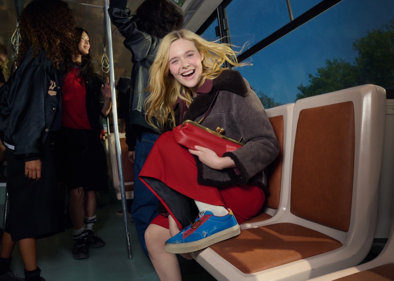 Elle Fanning smiling while seated on a bus holding a red clutch, wearing a shearling jacket and blue sneakers