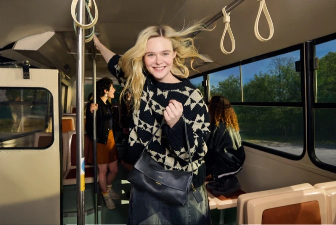 Elle Fanning holding a dark leather shoulder bag on a bus interior for the Coach Live Your Story campaign, photographed by Elaine Constantine
