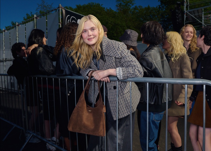 Elle Fanning leaning on a metal crowd barrier wearing a plaid blazer and holding a brown suede handbag.