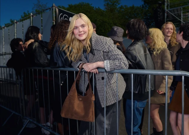 Elle Fanning leaning on a metal crowd barrier wearing a plaid blazer and holding a brown suede handbag.