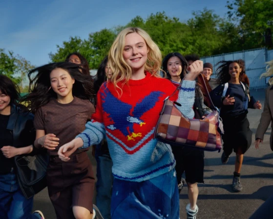 Elle Fanning leading a group of young adults running outdoors, wearing a red patterned sweater and holding a handbag for Coach.