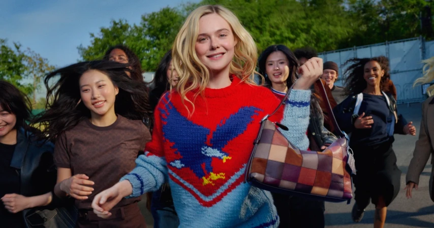 Elle Fanning holding a patchwork leather shoulder bag while running outdoors with a group of people, wearing a red and blue knitted sweater
