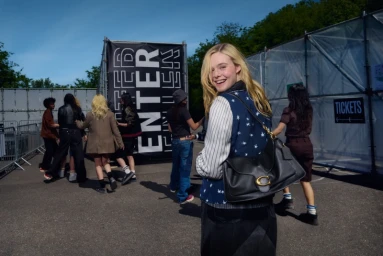 Elle Fanning smiling over her shoulder wearing a star-printed vest and carrying a black leather handbag outdoors for Coach.