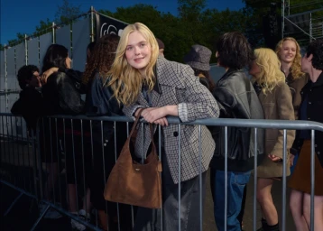 Elle Fanning leaning on a metal crowd barrier wearing a plaid blazer and holding a brown suede handbag.