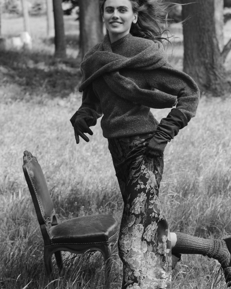 Black and white editorial image of a smiling woman wearing a knitted sweater and gloves, walking past an antique chair in a grassy meadow.