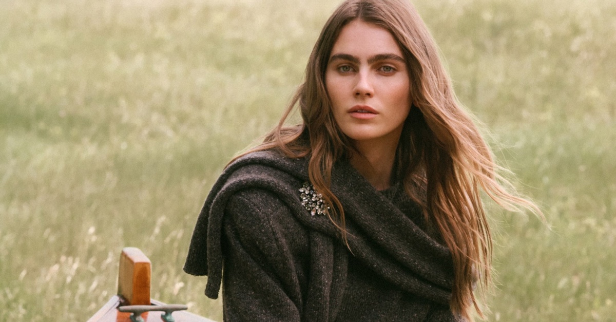 A woman wearing a dark wool coat with a jeweled brooch outdoors against a soft green meadow backdrop.