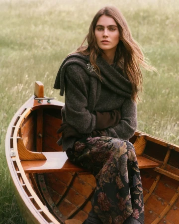 Woman sitting in a wooden boat wearing a dark grey sweater and floral patterned skirt, set in a grassy meadow.