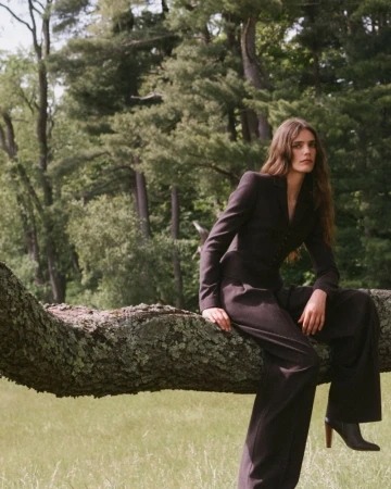Model wearing a dark tailored suit perched on a large mossy tree branch outdoors with a forest background