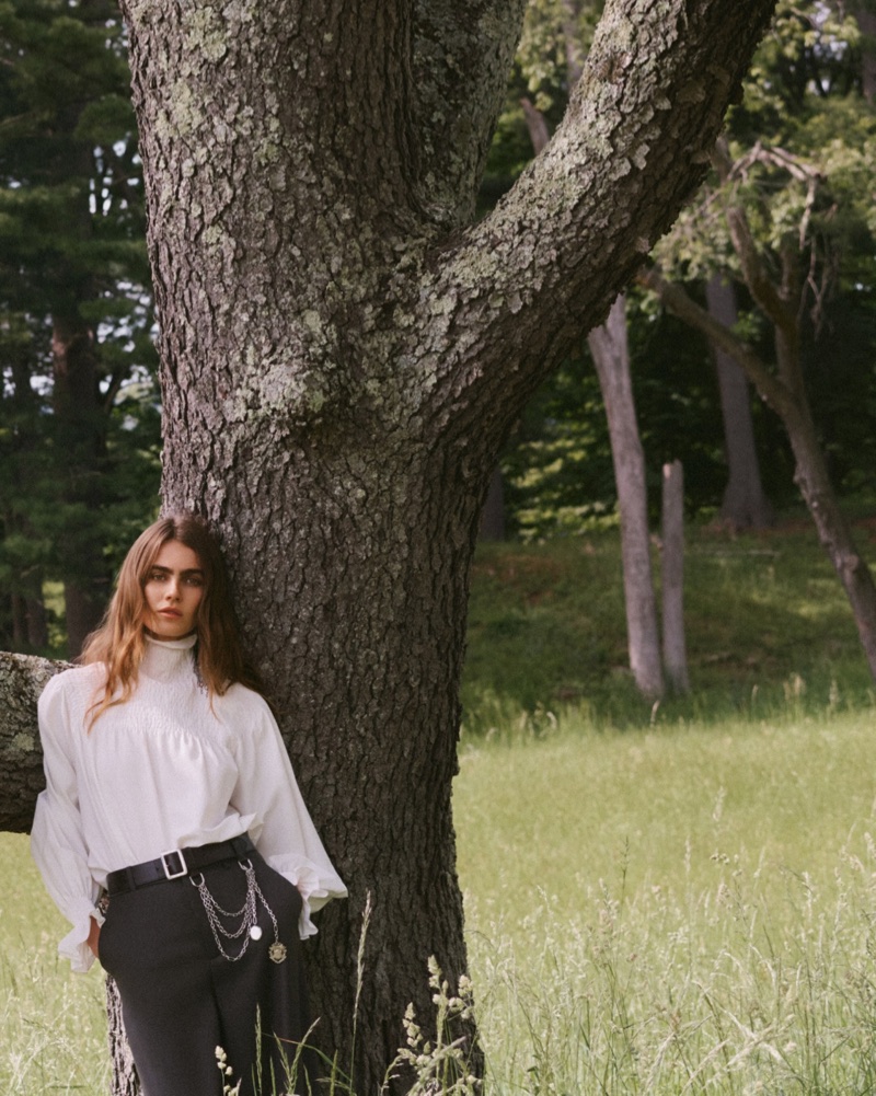 Model leaning against a large tree trunk in a grassy meadow wearing a white blouse and dark trousers with a chain belt.