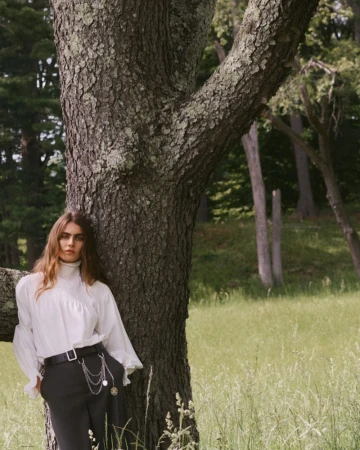 Model leaning against a large tree trunk in a grassy meadow wearing a white blouse and dark trousers with a chain belt.