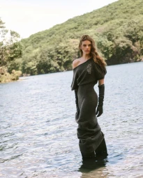 Model standing in a lake wearing a dark grey one-shoulder dress and long gloves with forested hills in background