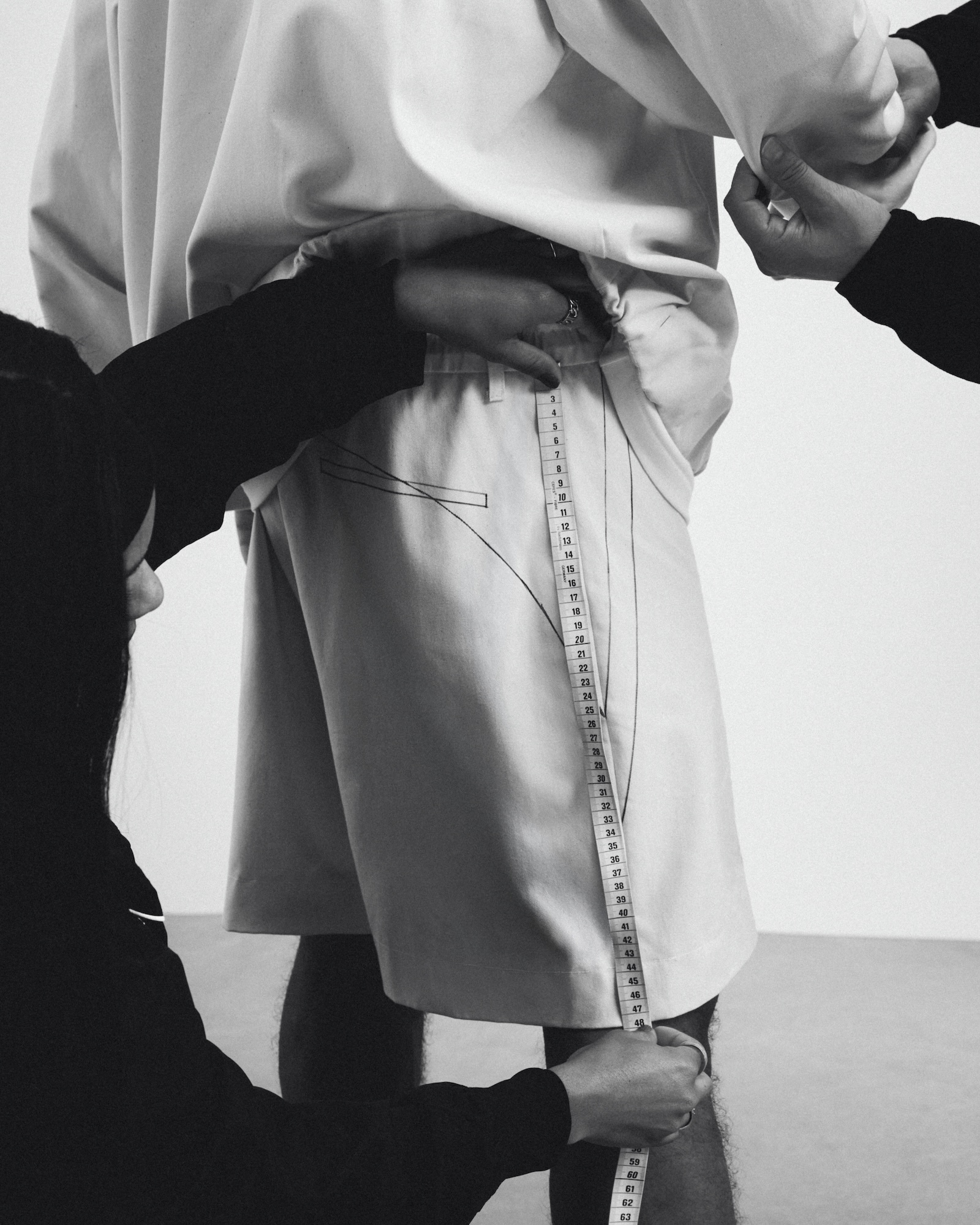 Tailor measuring shorts on a person standing against a plain white background in a monochrome studio setting