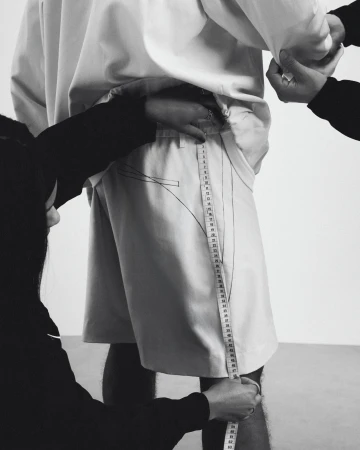 Tailor measuring shorts on a person standing against a plain white background in a monochrome studio setting