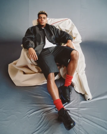Footballer Desire Doue seated in a makeshift fabric chair wearing a black jacket and red socks against a grey background