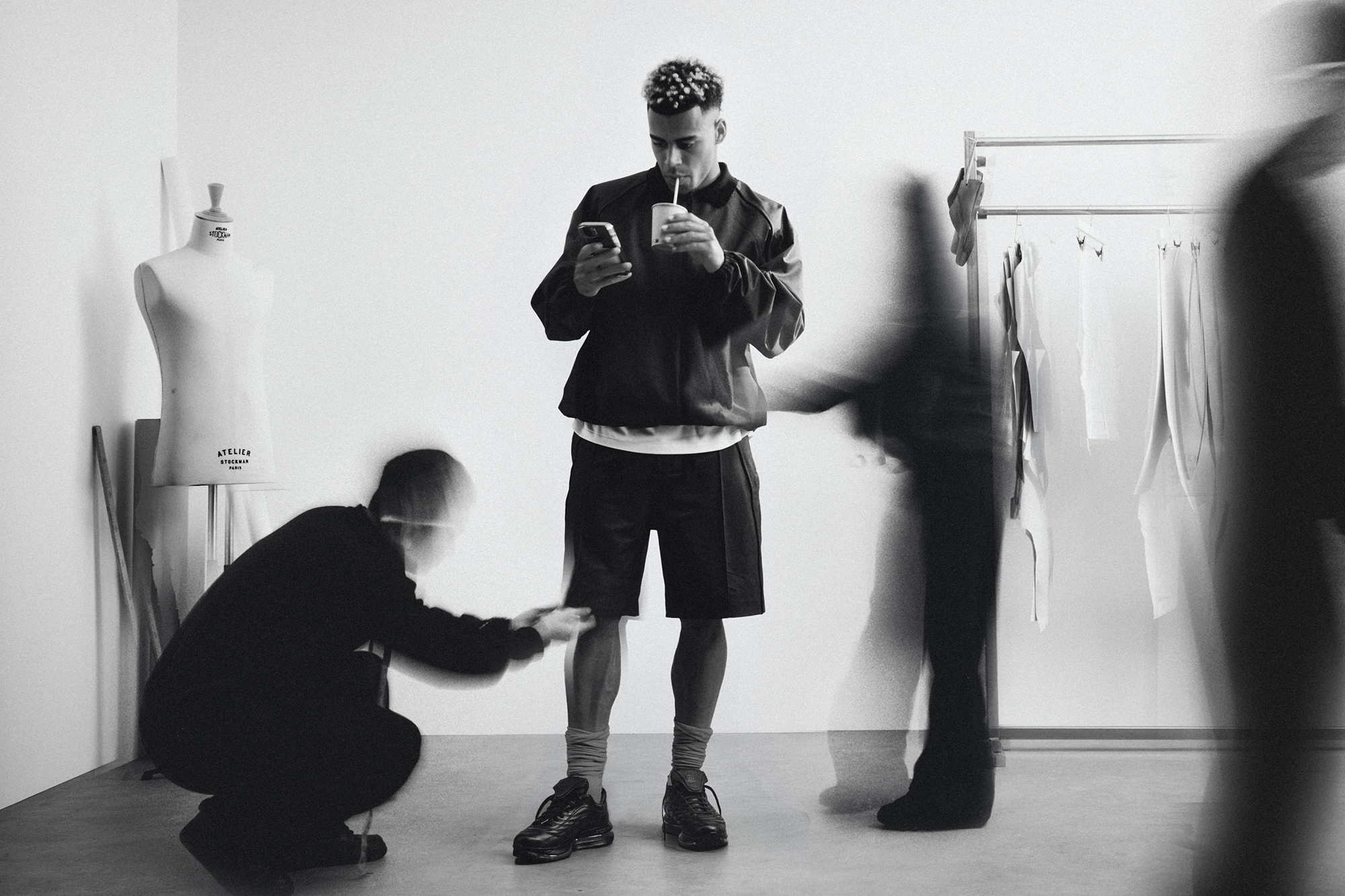 Black and white editorial portrait of footballer Desire Doue standing in a studio with blurred figures moving around him
