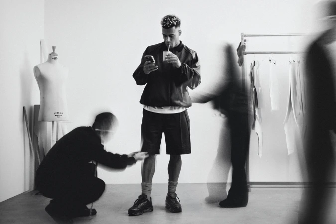 Black and white editorial portrait of footballer Desire Doue standing in a studio with blurred figures moving around him