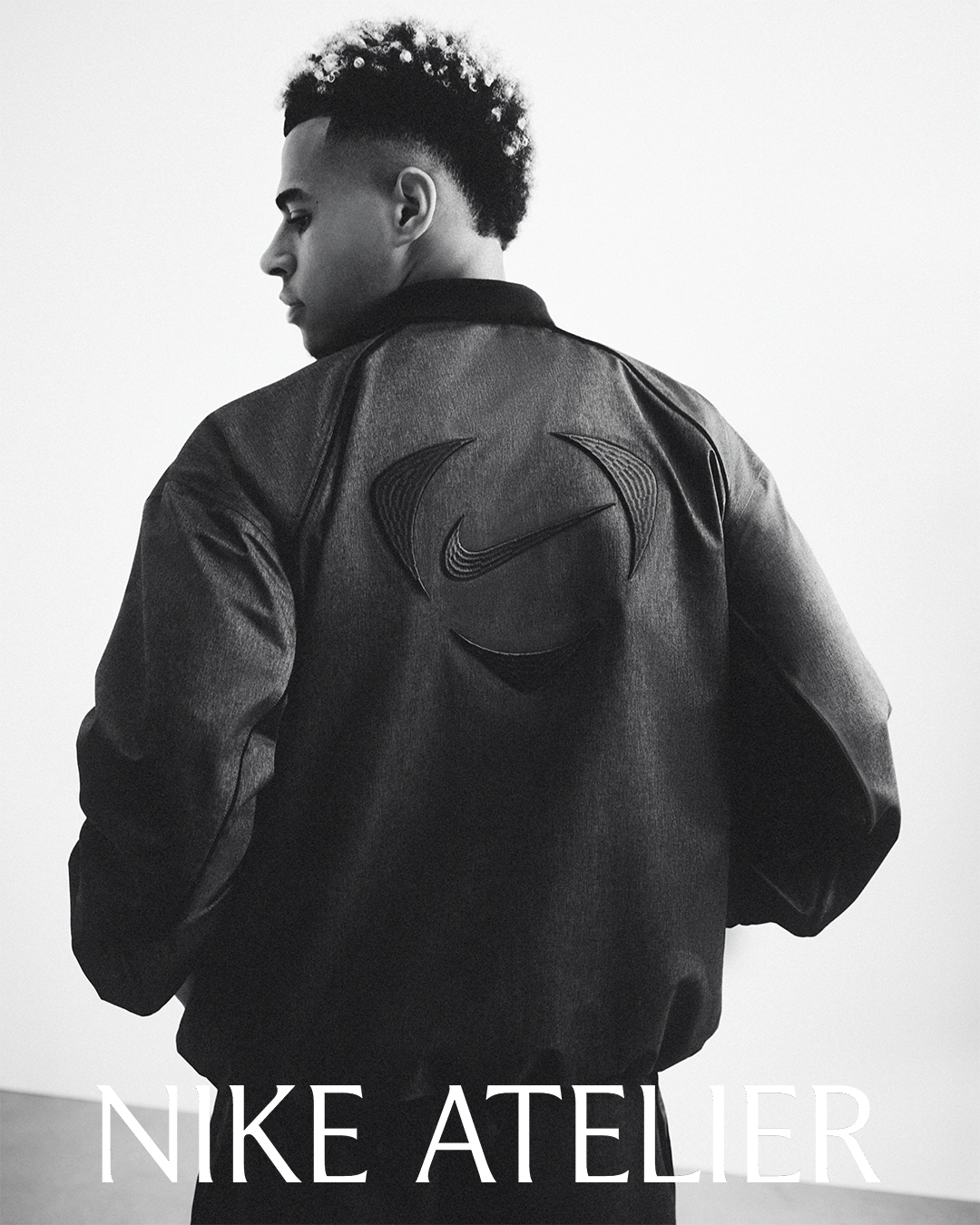 Footballer Desire Doue wearing a dark bomber jacket with embossed Nike branding against a white studio background.