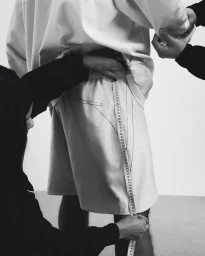 Tailor measuring shorts on a person standing against a plain white background in a monochrome studio setting