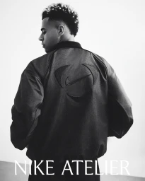 Footballer Desire Doue wearing a dark bomber jacket with embossed Nike branding against a white studio background.