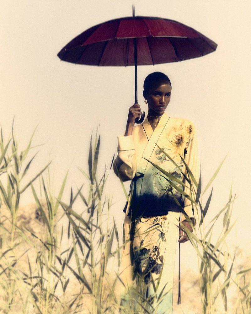 Adult woman in a floral kimono standing outdoors, holding a red umbrella in a field of tall green grass on a sunny day.
