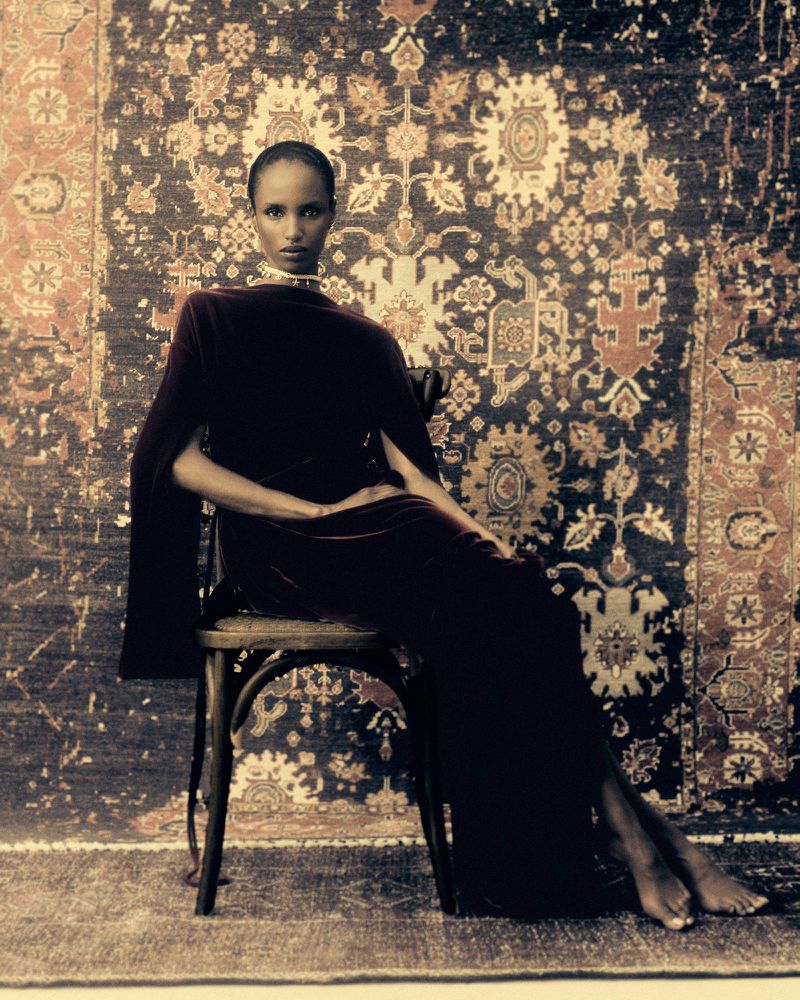 Elegant African woman in velvet gown sits on chair against ornate rug background in studio shot, fashion, beauty, and style editorial.