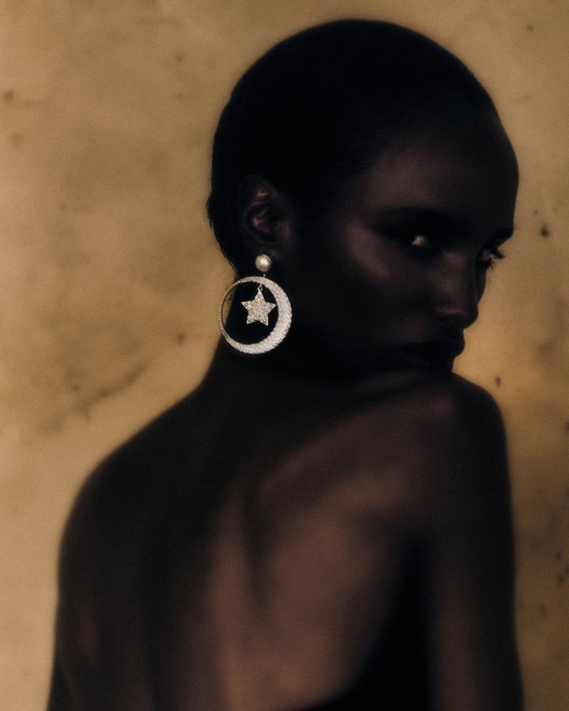 Elegant portrait of an African American woman with a bare shoulder, wearing a star and moon earring against a textured yellow backdrop.