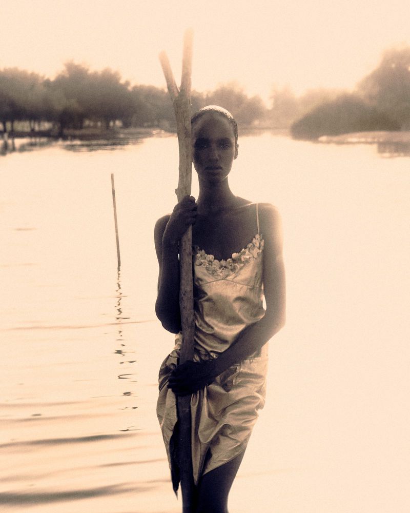 Woman in a shiny dress stands in water holding a stick with a backdrop of trees and bright sky, creating a serene and ethereal scene.